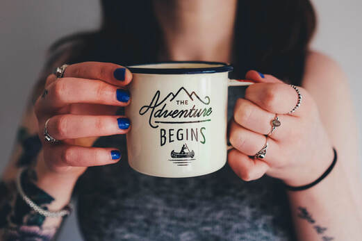 Picture of dark haired female with blue painted nails holding a coffee mug that reads The Adventure Begins